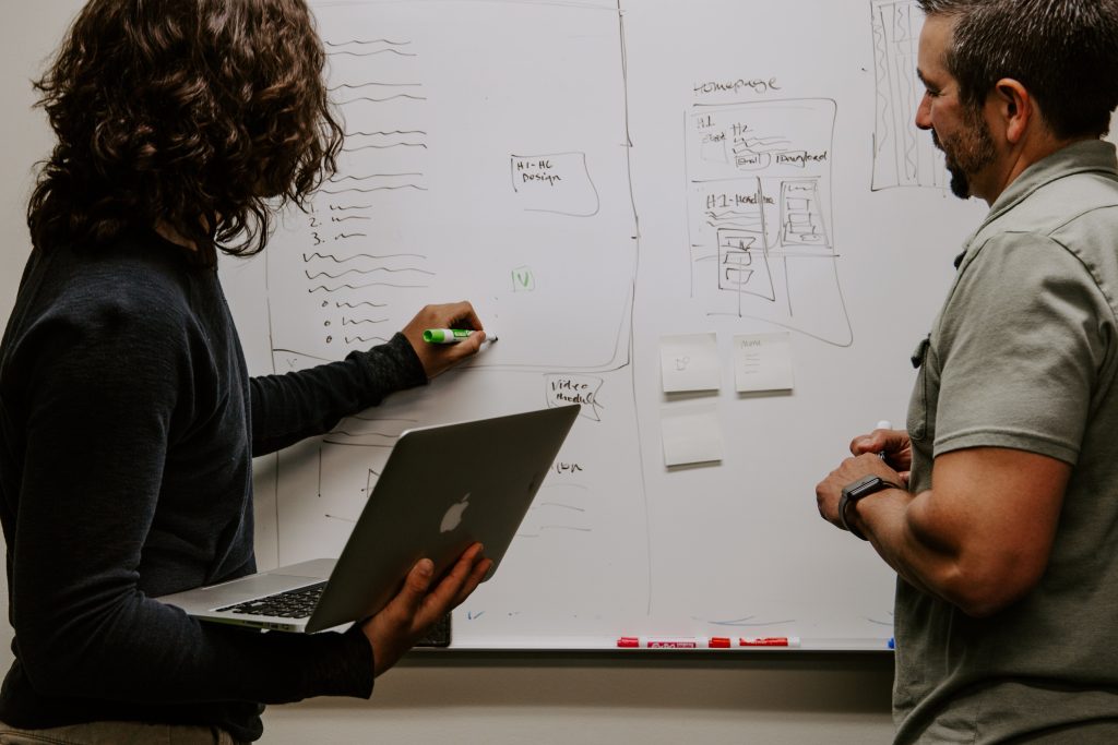 A woman holding a laptop writing on a whiteboard and a man observing the lecture.